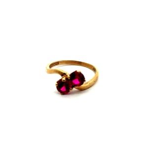 May include: A gold bypass ring featuring two round, vibrant pinkish-red gemstones set side-by-side. The band has a smooth, polished finish and curves gently to hold the stones in a bypass style, creating a continuous, flowing metal design.