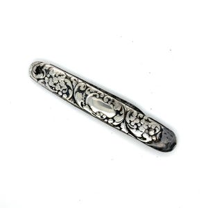 May include: A silver pocket knife with ornate floral and scrollwork engravings. The knife is closed, with a rounded end and a small hole for a lanyard. The design includes an oval central panel and detailed patterns.