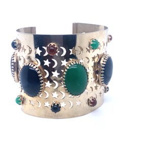 May include: A gold-toned cuff bracelet with cut-out star and crescent moon designs. The bracelet features large oval-shaped gemstones in black, green, and red. Small gemstones in matching colors are also set around the bracelet.