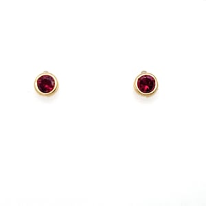 May include: A pair of stud earrings featuring round, deep red gemstones set in polished gold-toned metal bezels. The circular stones are securely held within the smooth, metallic frames, creating a simple and classic design for these small earrings.