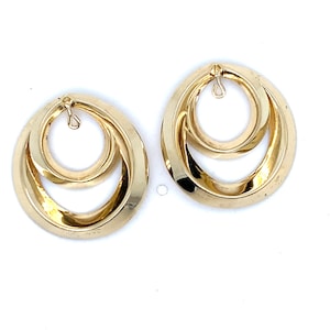 May include: A pair of gold-toned earrings featuring a double-hoop design. Each earring consists of a smaller circular hoop nested within a larger, slightly offset circular hoop. Both hoops have a smooth, polished finish with a small metal loop at the top.