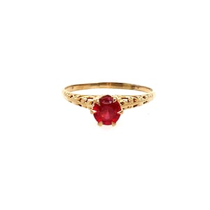 May include: A gold ring featuring a round, faceted red gemstone set in a six-prong mounting. The band is crafted with an intricate, openwork filigree design that extends from the setting along the sides of the ring, creating a detailed metalwork.