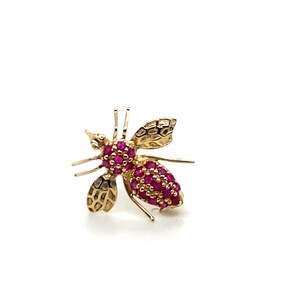 May include: A gold bee-shaped brooch featuring a head and abdomen encrusted with small, vibrant pink gemstones. The insect has textured, polished gold wings and thin gold legs extending from the central body, creating a detailed, insect-inspired design.