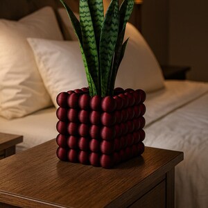May include: A decorative, dark red planter with a unique, textured design, filled with a green snake plant. The planter is square-shaped and sits on a wooden nightstand. The plant's leaves have a distinctive pattern.