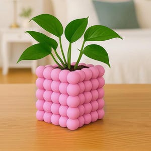 May include: A square, pink, bubble-textured planter filled with a green leafy plant. The planter sits on a wooden surface. The background includes a bed with white pillows and a framed picture.