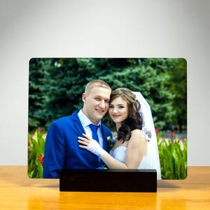 May include: A rectangular photo print featuring a smiling couple, displayed on a wooden stand. The couple is dressed in wedding attire, with the man in a blue suit and the woman in a white dress and veil. The background shows greenery.
