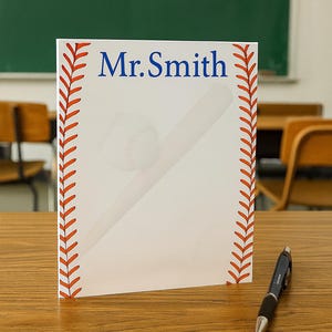 May include: A white notepad with a baseball theme, featuring a baseball and bat graphic. The notepad has the name "Mr. Smith" in blue text and a red baseball stitch design along the edges. A pen rests on the wooden desk.