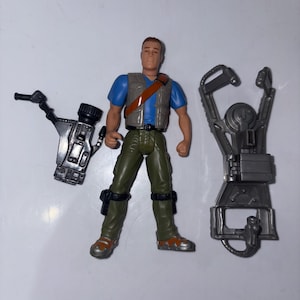 Puede incluir: Una figura de acción de plástico de un hombre con una camisa azul, pantalones verdes y un chaleco marrón. La figura tiene cabello castaño y sostiene una correa marrón. Dos accesorios grises están al lado de la figura.