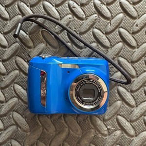Puede incluir: Una cámara digital Kodak azul brillante con una correa negra. La cámara tiene una lente plateada y el texto "KODAK AF 3x Optical Aspheric Lens" es visible. La lente tiene un rango de 34mm - 102mm (Equivalent).