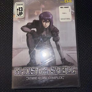 以下が含まれることがあります： 「攻殻機動隊 STAND ALONE COMPLEX」Vol. 04のDVDケース。黒い衣装のキャラクターがひざまずいているカバー。タイトルは銀色の未来的なフォントです。「Suggested 13 Up」ステッカーが左上にあります。