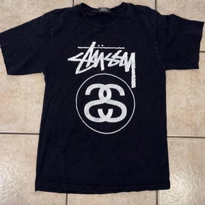 May include: Black t-shirt featuring the white "Stüssy" logo and a double-S circle graphic. The short-sleeved shirt is made of a soft material, suitable for casual wear.