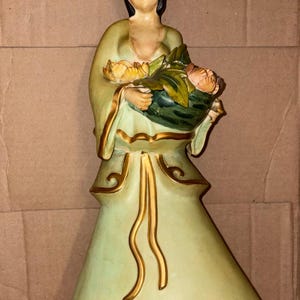 Könnte beinhalten: Keramikfigur einer Frau in einem fließenden grünen Kleid, die einen Blumenkorb hält. Die Figur hat einen ruhigen Ausdruck und ist mit goldenen Akzenten verziert. Die Blumen sind eine Mischung aus Gelb und Pfirsich.