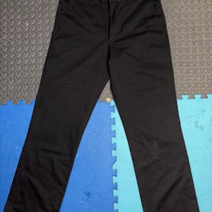 May include: Black trousers with a button and zipper closure. The pants have a straight leg design and a waistband with belt loops. The fabric appears to be a sturdy material, suitable for casual or work wear. The pants are laid flat on a patterned surface.