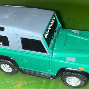 Puede incluir: Un juguete verde y gris de un Defender, con neumáticos negros y una rueda de repuesto en la parte trasera. El texto "DEFENDER" está en el parabrisas y "4x4" en la ventanilla lateral. El juguete está sobre una superficie verde.