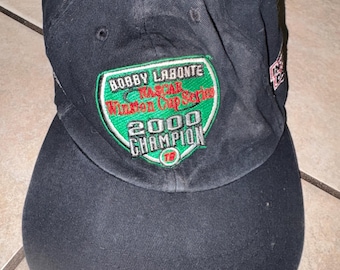 May include: A black baseball cap featuring an embroidered patch on the front. The patch is green with a black border and white text that reads Bobby Labonte NASCAR Winston Cup Series 2000 Champion, with the number 18 in a red circle.
