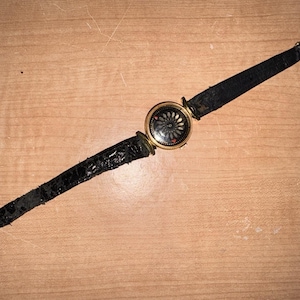 Puede incluir: Un reloj de pulsera antiguo con una caja dorada y una correa de cuero negro. La esfera del reloj tiene un diseño oscuro con un patrón floral. La correa muestra signos de desgaste.
