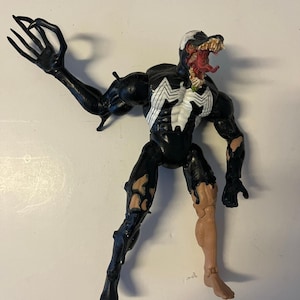 May include: A black and white Venom action figure with a detailed design. The figure has a white spider emblem on the chest, a gaping mouth with a red tongue, and a claw-like hand. The figure is approximately 6 inches tall.