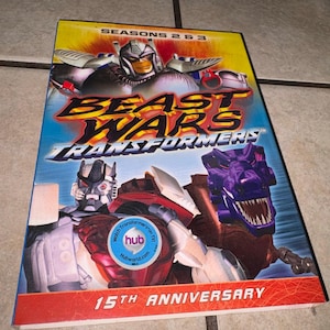 Puede incluir: Una caja de DVD de la serie animada "Beast Wars: Transformers". La portada presenta personajes robot en rojo, plateado y morado, con el texto "Temporadas 2 y 3" y "15th Anniversary". También se ve un logotipo de Hub Network.