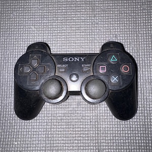 May include: Black Sony PlayStation 3 controller with directional pad, action buttons, and analog sticks. The controller has the Sony logo and Select and Start buttons. The background is a textured gray surface.