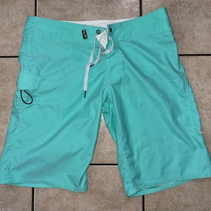May include: Turquoise board shorts with a white drawstring and a small side pocket. The shorts have a white waistband and are made of a lightweight material. The shorts are designed for water activities.