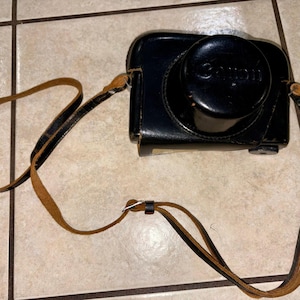 May include: Black leather camera case with a brown leather strap. The camera case has the brand name "Canon" embossed on the top. The strap is attached to the case with metal hardware.