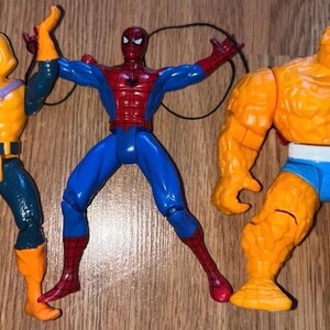 以下が含まれることがあります： コミックキャラクターのアクションフィギュア3体。フィギュアには、黄色とオレンジ色のゴブリン、赤と青のスパイダーマン、オレンジ色のシングが含まれます。フィギュアは木製の表面に配置されています。