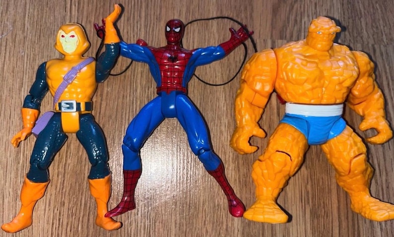 以下が含まれることがあります： コミックキャラクターのアクションフィギュア3体。フィギュアには、黄色とオレンジ色のゴブリン、赤と青のスパイダーマン、オレンジ色のシングが含まれます。フィギュアは木製の表面に配置されています。
