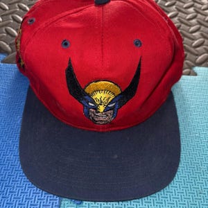 May include: A red baseball cap with a navy blue brim. The cap features a Wolverine character design on the front. The design includes a yellow, blue, and black color scheme. The cap has a button on top.