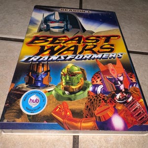 Op de afbeelding: Een dvd-hoes voor "Beast Wars: Transformers" Seizoen 1. De cover toont illustraties van robotkarakters in heldere kleuren, met de titel van de show in vette, gestileerde letters. Een ronde sticker leest "Watch Transformers on TV!" en "hubworld.com".