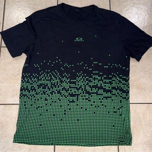May include: Black short-sleeved t-shirt with a green dot pattern that fades from top to bottom. The Oakley logo is centered on the chest. The shirt is made of a lightweight, breathable material.