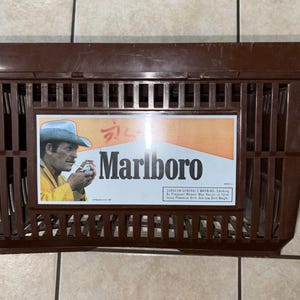 May include: A brown plastic basket with a rectangular shape. The basket features a rectangular graphic with the Marlboro logo and an image of a man in a cowboy hat. The basket has a grid-like design.