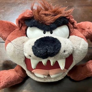 May include: A plush toy of the Tasmanian Devil, featuring a red-brown fur, large white teeth, and a black nose. The toy has a wide, open mouth and is lying on a wooden surface.