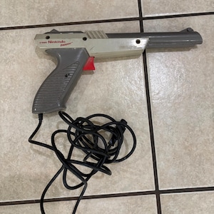 May include: A grey and off-white Nintendo Zapper light gun controller with a red trigger. The device features a long barrel, a textured grip, and a black coiled cord extending from the base. The text Nintendo Zapper is printed on the side.
