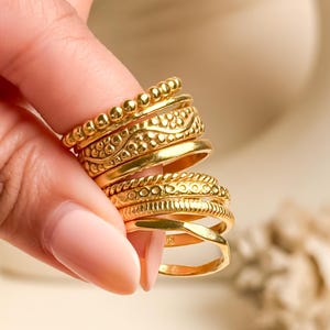 Peut inclure: Plusieurs bagues dorées aux motifs détaillés, dont des motifs perlés et texturés. Les anneaux sont empilés, mettant en valeur leurs différents styles. Les bijoux sont présentés sur un fond doux et neutre.
