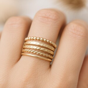 May include: A selection of gold rings displayed on a finger. The rings feature diverse designs, such as beaded, hammered, twisted, and plain bands. The rings are crafted from a bright, yellow gold material.