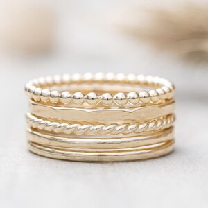 May include: A collection of gold-coloured rings. The rings feature diverse designs, including a beaded band, a hammered band, a twisted rope band, and smooth bands. The rings are stacked, creating a layered effect.