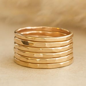May include: A collection of six gold rings, each with a hammered texture. The rings are stacked together, displaying their similar design and warm, metallic colour. The hammered effect gives a faceted appearance. The background is a soft, neutral tone.