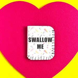 May include: A small, rectangular object with the text "SWALLOW ME" printed on it, set against a vibrant pink heart shape on a yellow background. The object has a white base with a colorful geometric pattern.