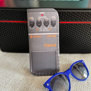 Peut inclure: Une pédale de guitare noire et grise avec le mot "Metal" imprimé dessus. La pédale a des boutons étiquetés "Check", "Level", "Equalizer" et "Dist".
