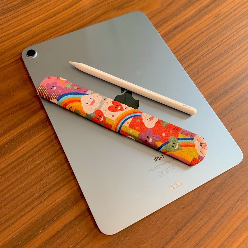 May include: A silver iPad with a white Apple Pencil and a colorful fabric sleeve with a rainbow and heart design. The iPad has the word "iPad" printed on the back.