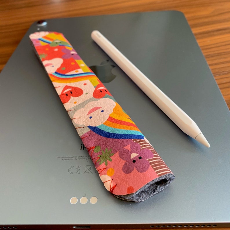 May include: A colorful patterned pencil holder with a gray felt lining. The holder is designed to fit an Apple Pencil and is lying on top of a silver iPad.
