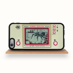 May include: A phone case with a retro handheld game design. The case is beige with a black border and features a screen displaying an octopus and ship scene. The text on the case includes "TAЙHЫ OKEAHА" and "ЭЛЕКТРОНИКА". Red buttons and accents complete the design.