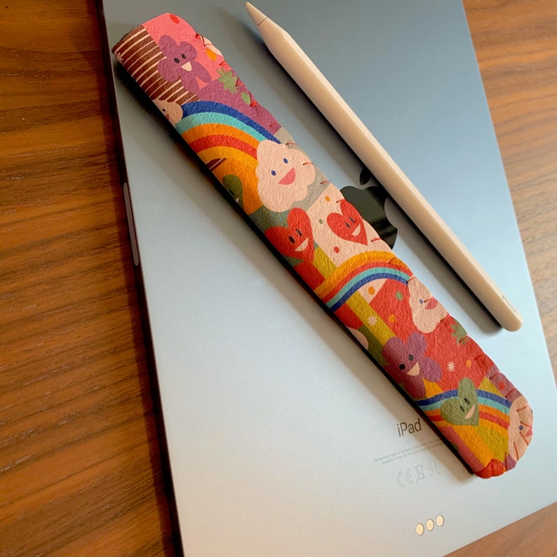May include: A colorful fabric pencil grip with a rainbow, hearts, and clouds design. The grip is on top of a silver iPad with the word "iPad" printed on the back.