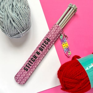 May include: A pink knitting needle case with the text "STITCH'N BITCH" in black. The case holds three gray knitting needles and is accessorized with a colorful charm. A ball of gray yarn and a ball of red yarn are also in the image.