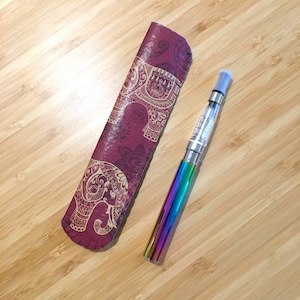 May include: A burgundy case with gold elephant designs next to a rainbow-coloured vape pen. The case is made of a textured material. The vape pen has a clear tank and a blue mouthpiece. The items are on a light brown wooden surface.