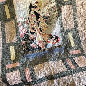 Könnte beinhalten: Eine gesteppte Decke mit einem zentralen Bild einer Geisha in einem Kimono, umgeben von floralen Mustern. Die Steppdecke hat ein rosa-graues Farbschema mit rechteckigen und gestreiften Akzenten. Die Geisha ist mit komplizierten Details und leuchtenden Farben dargestellt.