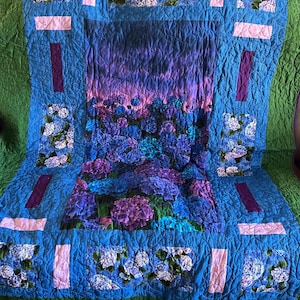 May include: A quilted blanket with a floral design. The blanket features a central panel of blue and purple flowers, framed by a blue border with pink and purple accents. The edges are green.