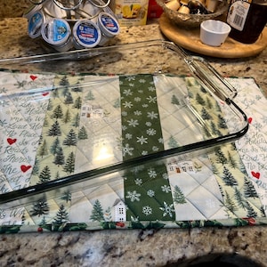 Puede incluir: Una fuente para hornear de vidrio transparente descansa sobre una almohadilla térmica acolchada con temática navideña. La almohadilla presenta un diseño de patchwork con acentos verdes, blancos y rojos, incluyendo texto, copos de nieve y árboles de Navidad.