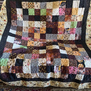 Puede incluir: Un edredón de retazos con una variedad de cuadrados estampados en tonos de marrón, rosa, verde y negro. El edredón tiene un borde negro y un reverso color crema con un patrón repetido de búhos y gatos.