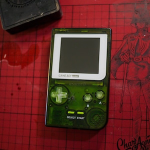 May include: A transparent green Game Boy Pocket handheld console. The device has a black screen, white border, and green buttons. The text "GAME BOY pocket" is visible above the screen, and "SELECT START" is printed on the bottom.
